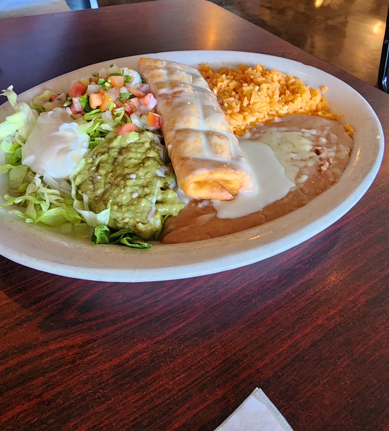 A rolled chimichanga under white cheese sauce with guacamole, sour cream, pico de gallo, rice and refried beans