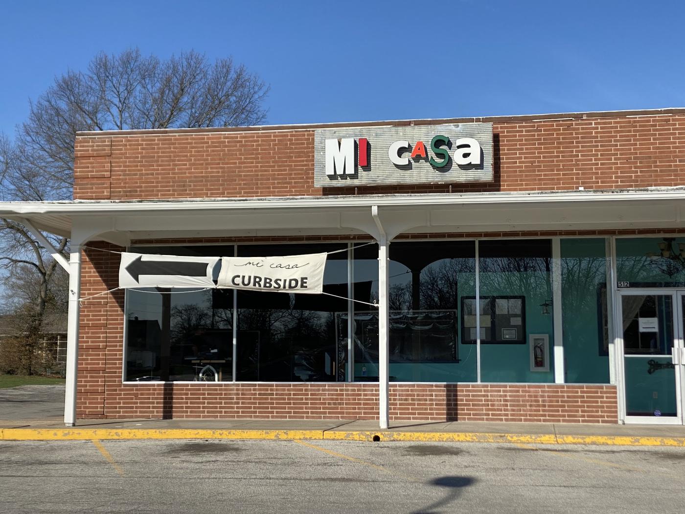 The Mi Casa storefront in daylight, brick front with the corrugated MI CASA sign above and a curbside banner across the window